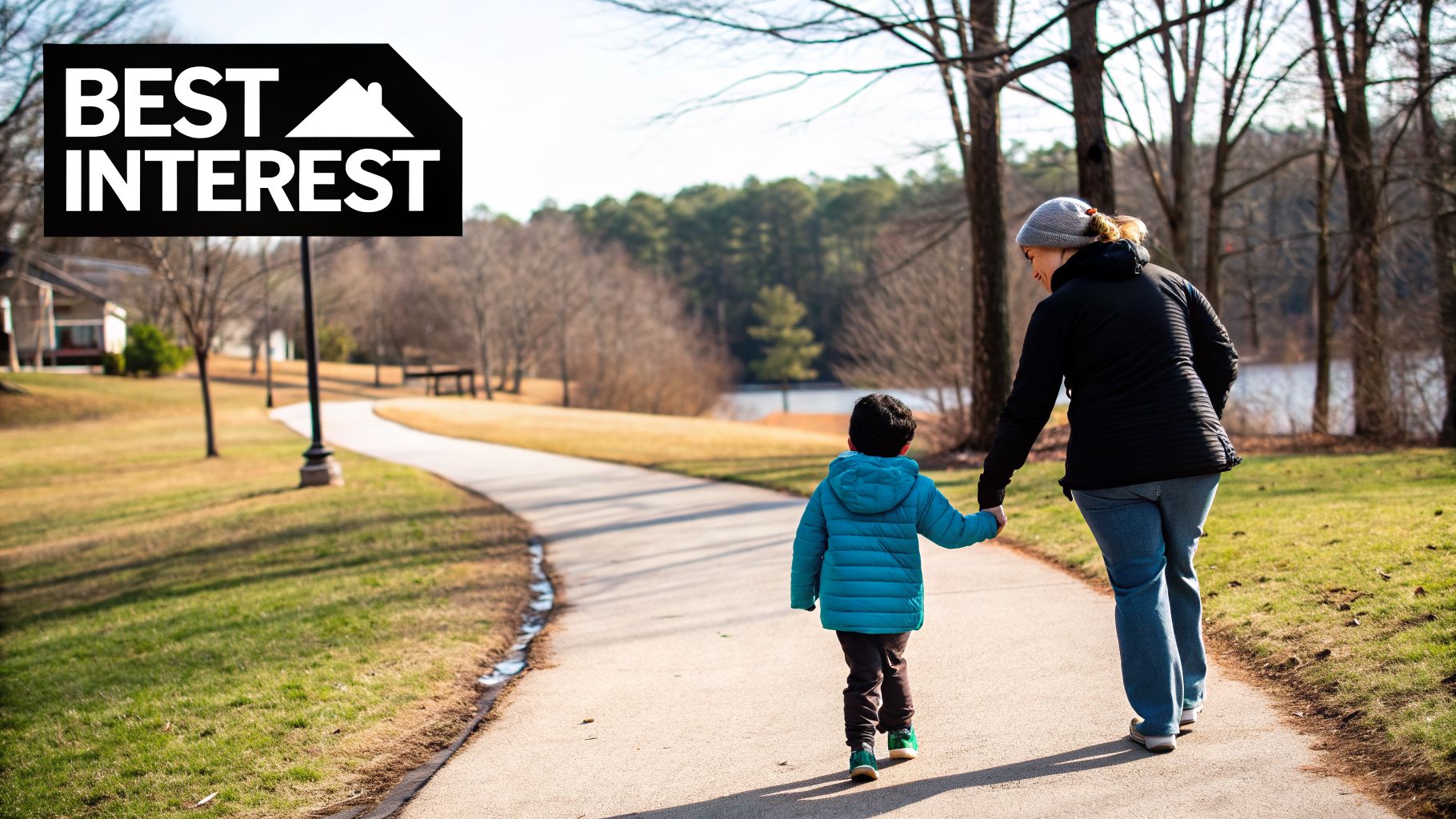 A woman and child hold hands, walking on a park path surrounded by trees and a lake.