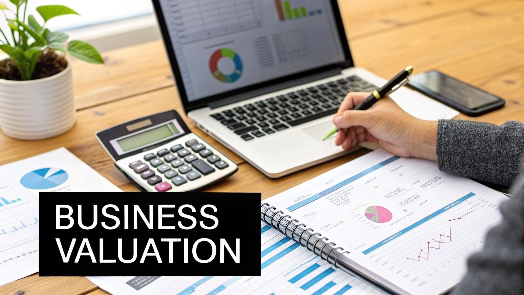 A person analyzes business valuation documents, graphs, and data using a laptop and calculator on a wooden desk.