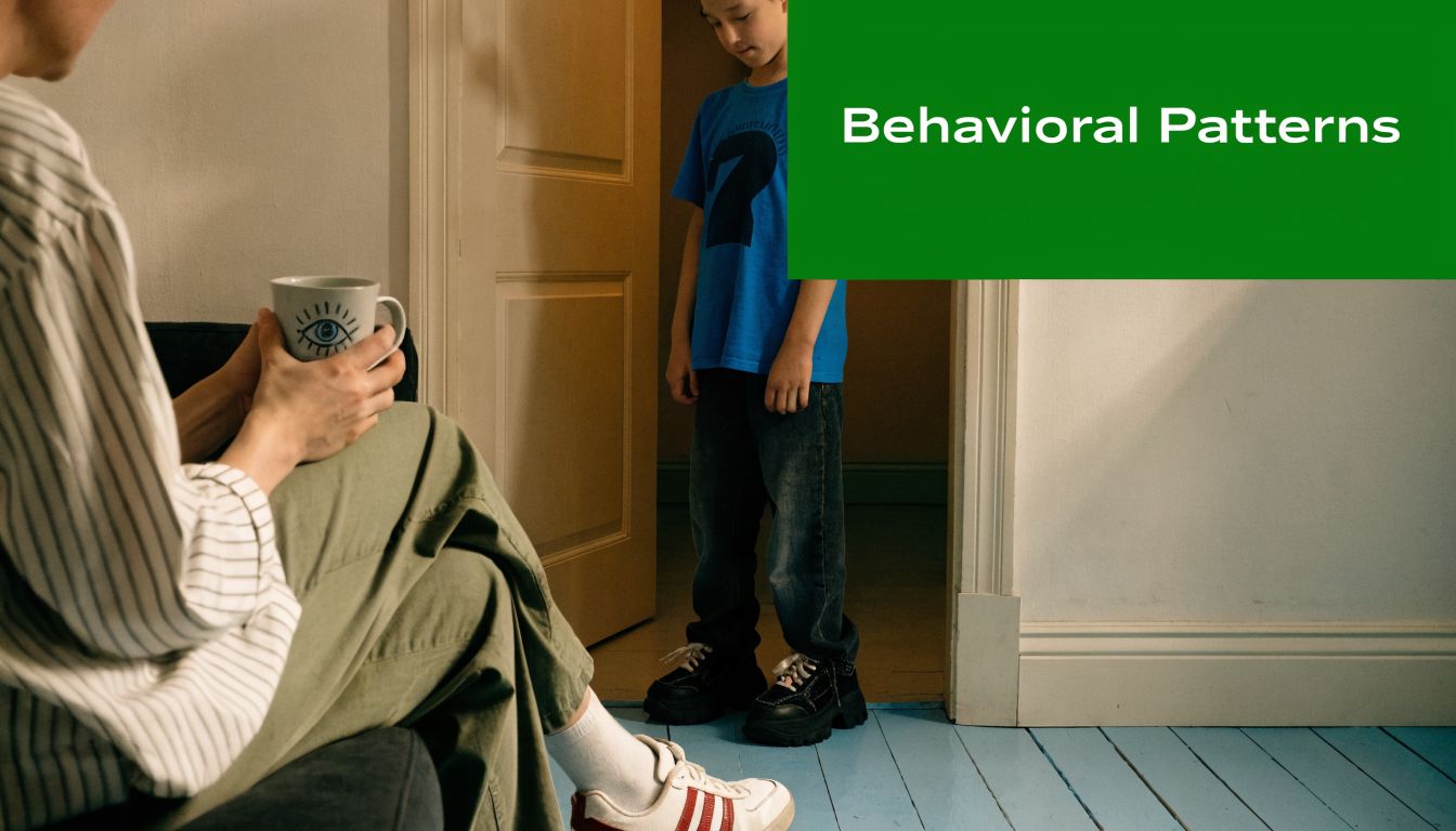 A young boy stands quietly in a doorway while a parent watches him from a couch.