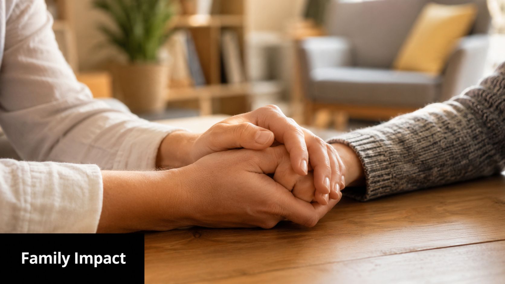 Two people holding hands across a wooden table, representing support, comfort, and family impact.